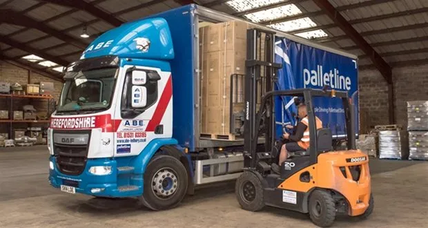 Forklift driver loading a large wooden crate into an ABE Palletline trailer inside a warehouse.