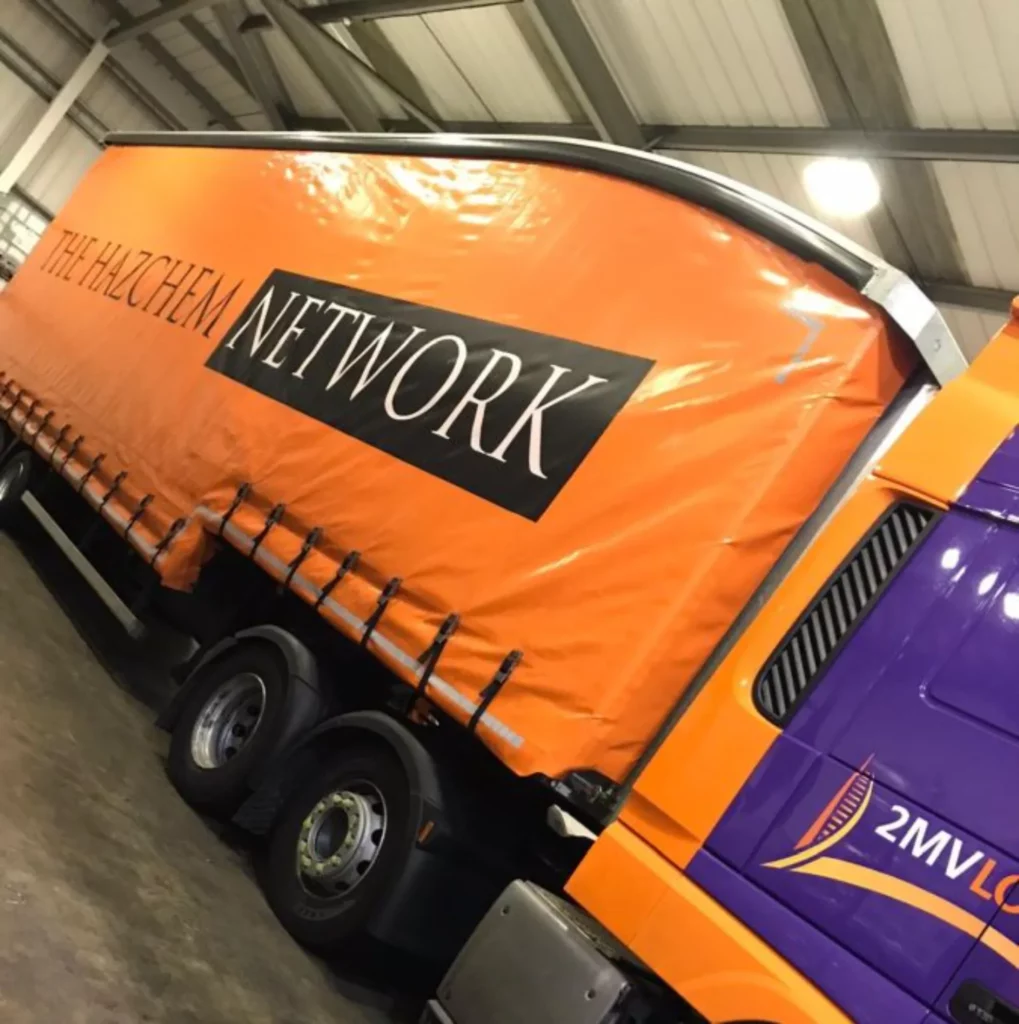 A brightly coloured 2MV Logistics lorry featuring an orange trailer with “The Hazchem Network” branding and a purple cab parked inside a warehouse facility.