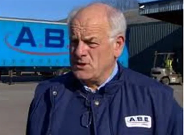 Older man in an ABE jacket speaking outdoors, with a blue ABE trailer and forklift in the background